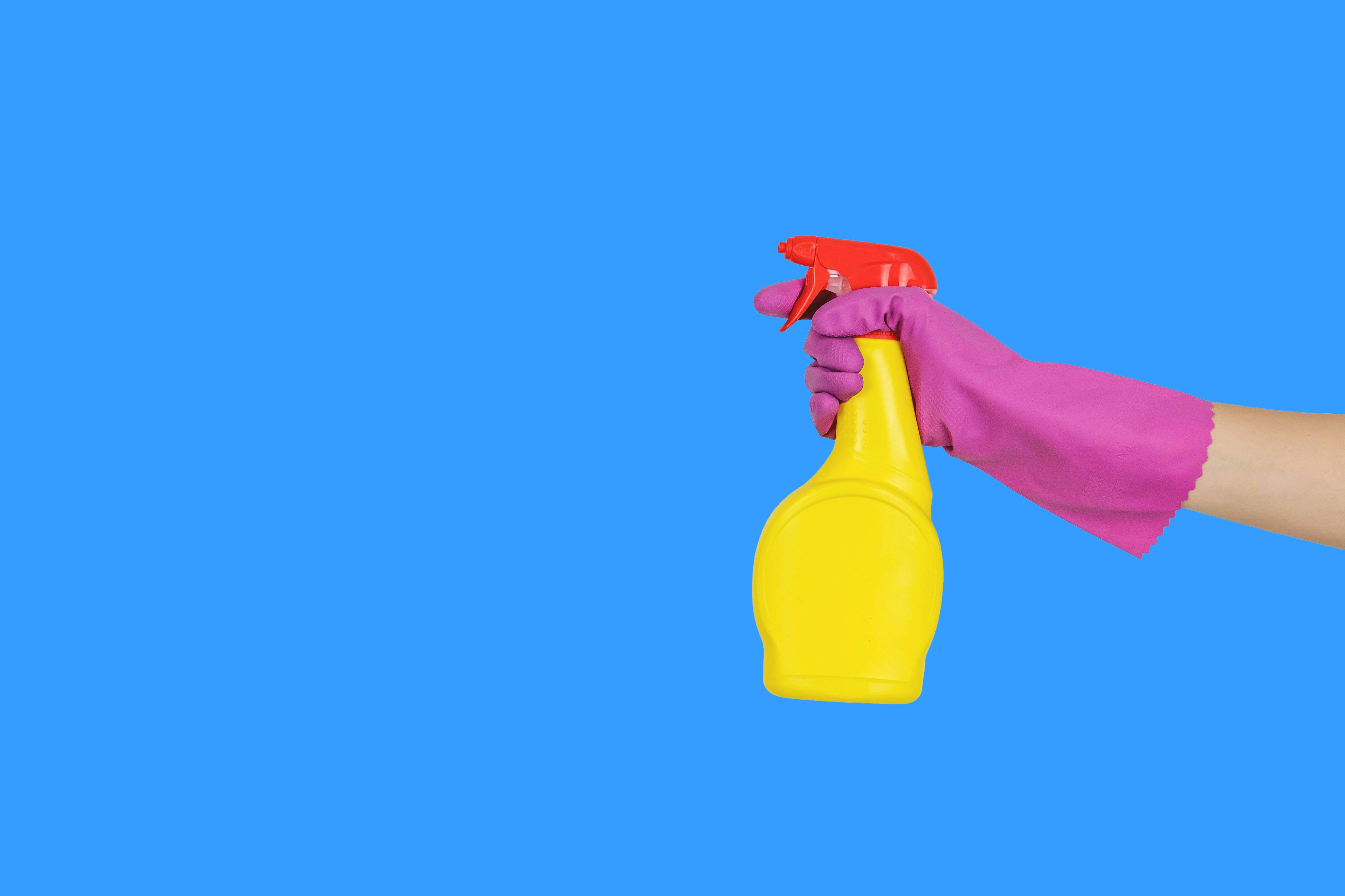 Hand wearing pink gloves holding a yellow cleaning spray bottle on a clean blue background for PrimeVita hero banner