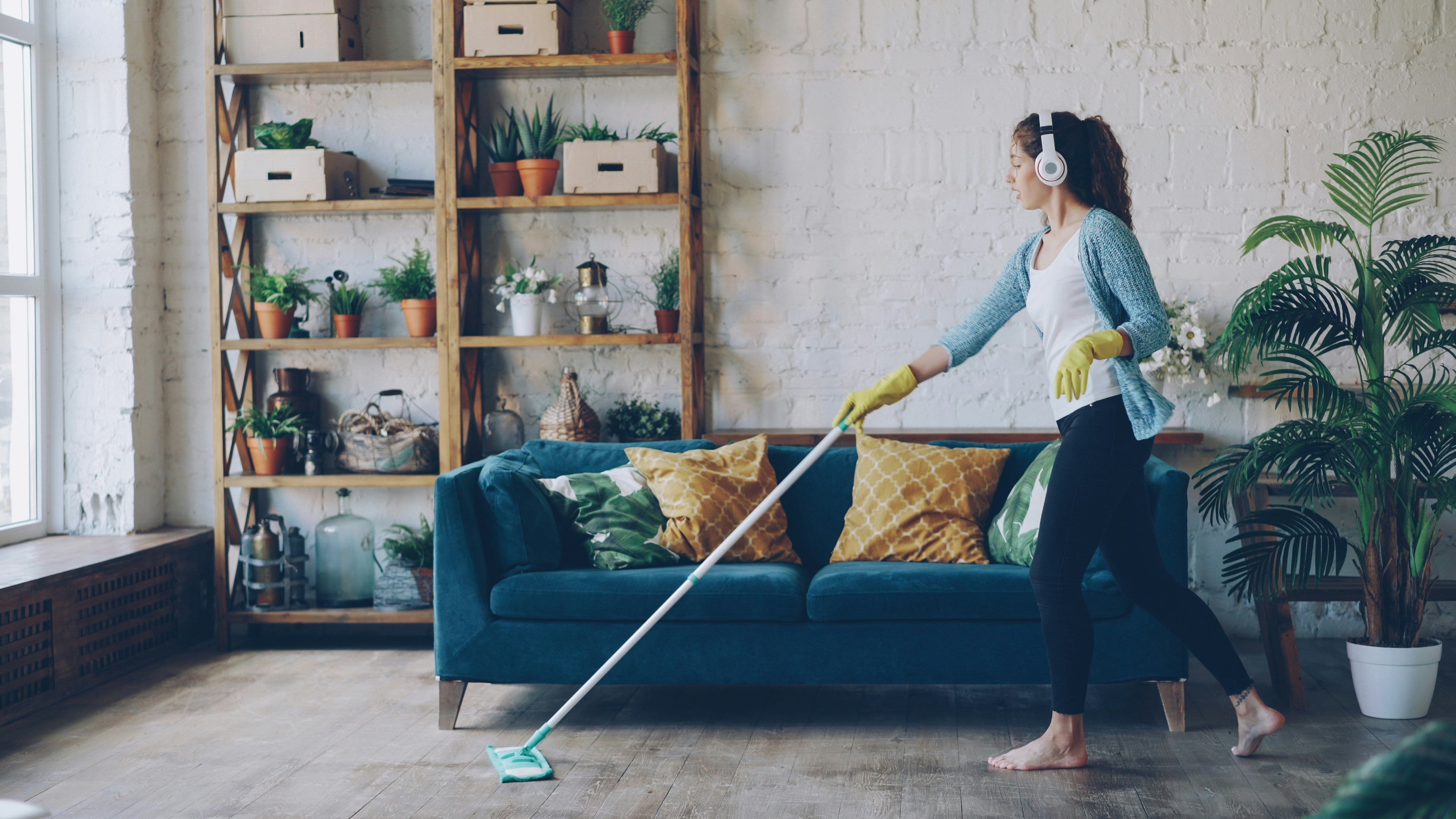 Woman wearing gloves and headphones mopping the living room floor in a bright, modern home with plants and organized shelves